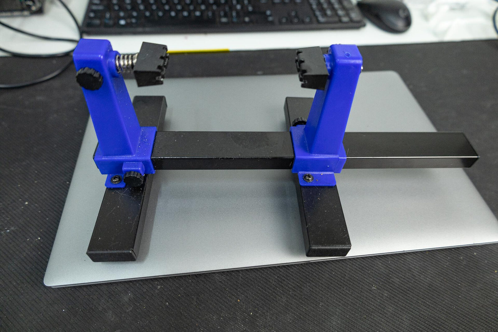 An adjustable blue and black PCB holder with sprung jaws, sitting on a workbench