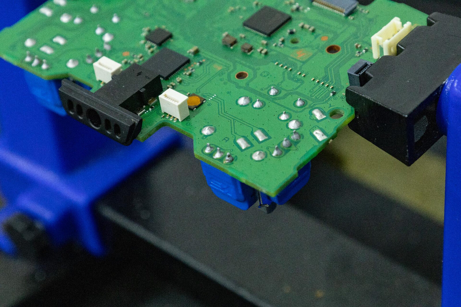 The underside of a DualSense board held in a PCB holder, showing bright conical solder joints on the joystick module pins