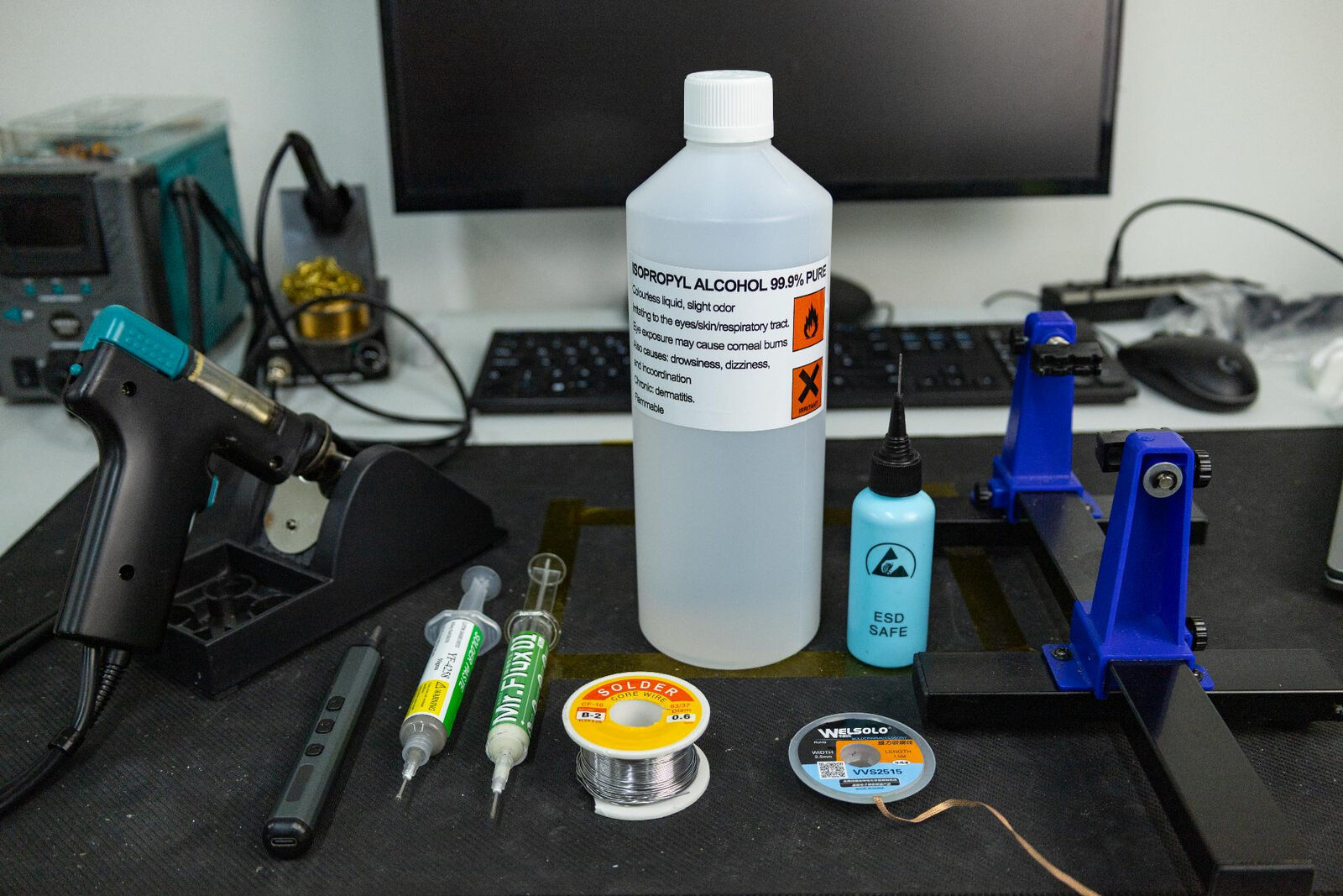A workbench laid out with a desoldering gun, isopropyl alcohol, flux and solder paste syringes, 63/37 solder, solder braid and a PCB holder